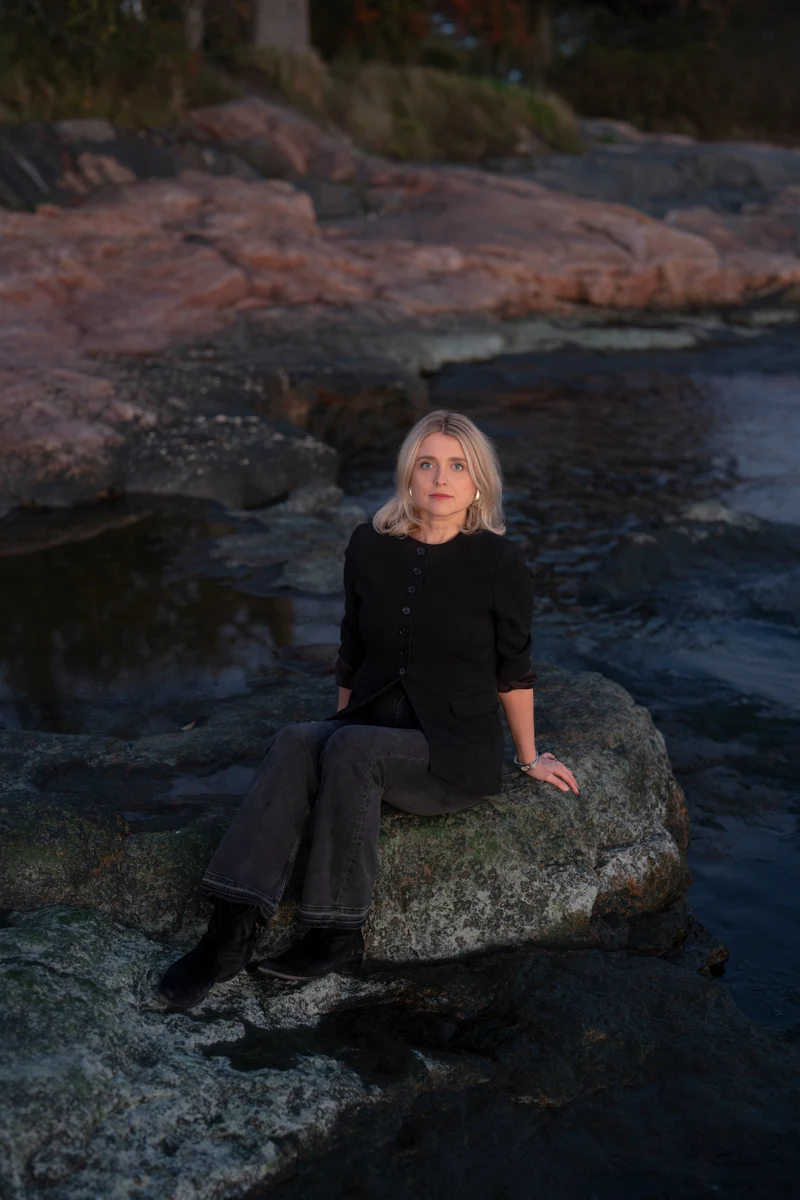 Joanna Numminen, founder of Numminen Advisory, seated on a rock on the Helsinki shoreline at dusk